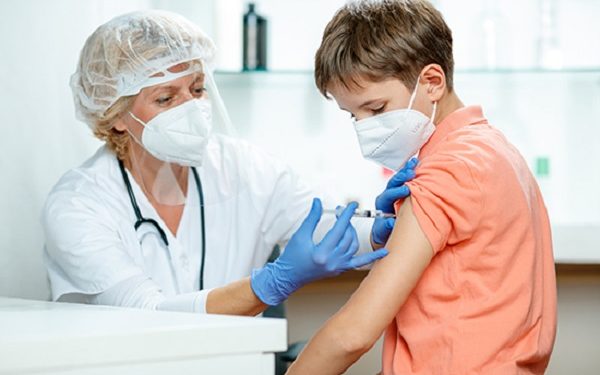 Female doctor fully equipped with protective workwear injecting vaccine into boy's arm, boy wearing protective face mask